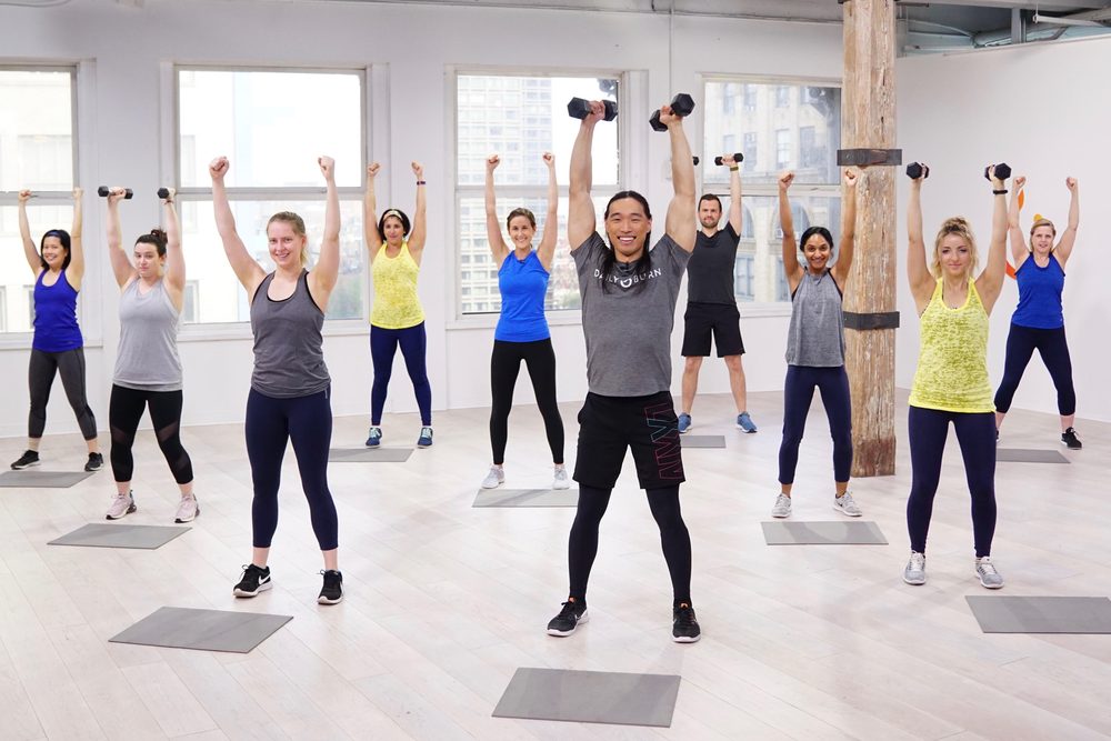 Chris leading a full studio class, dumbbells overhead